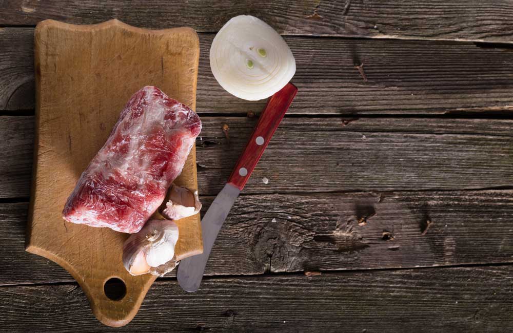 frozen steak on cutting board with knife, garlic and onion