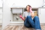 woman sitting on floor in front of leaky sink with wrench in one hand while talking on phone
