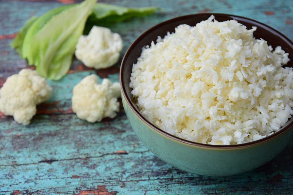 Cauliflower Rice in a bowl on a blue table with cauliflower flowerettes