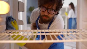 man cleaning the inside of a fridge looking like he wants to be somewhere else