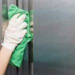 Gloved hand cleaning front of stainless steel fridge with a green rag