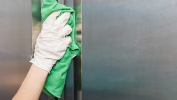 Gloved hand cleaning front of stainless steel fridge with a green rag