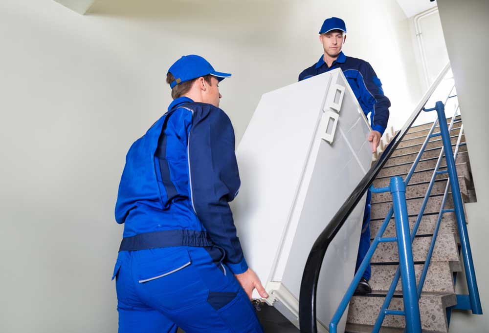 2 men in blue jumpsuits moving a refrigerator up stairs