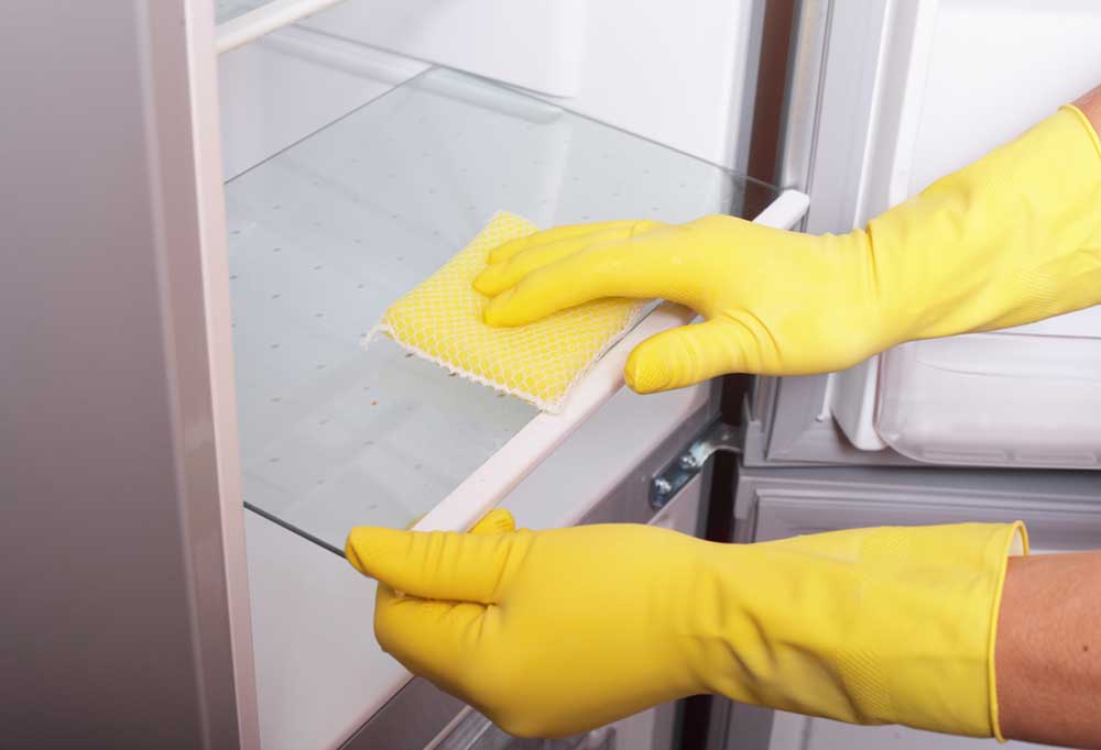 Yellow gloved hands with a sponge cleaning glass fridge shelves