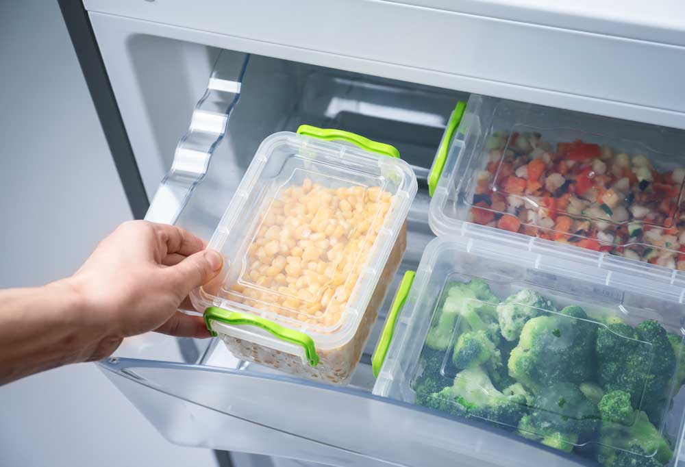 A hand reaching into a clear kitchen drawer to remove food storage containers of corn, broccoli, and mixed vegetables.