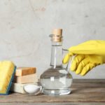 Yellow gloved hand holding the handle on a bottle of vinegar next to 3 bars of soap, baking soda in a bowl, an a yellow and blue sponge, all on a wooden table.