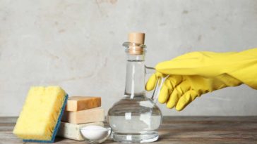 Yellow gloved hand holding the handle on a bottle of vinegar next to 3 bars of soap, baking soda in a bowl, an a yellow and blue sponge, all on a wooden table.