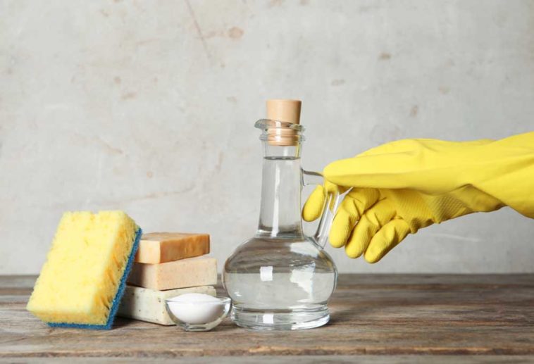 Yellow gloved hand holding the handle on a bottle of vinegar next to 3 bars of soap, baking soda in a bowl, an a yellow and blue sponge, all on a wooden table.