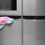 Hand in a pink glove wiping a stainless steel fridge with a blue rag