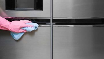 Hand in a pink glove wiping a stainless steel fridge with a blue rag
