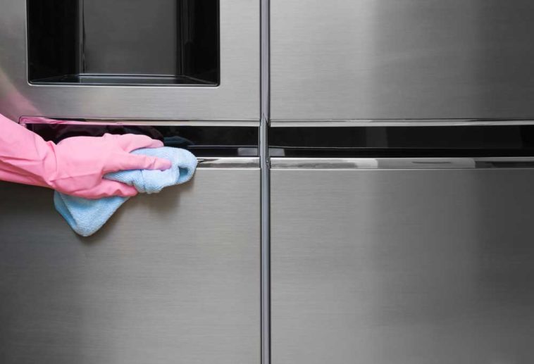 Hand in a pink glove wiping a stainless steel fridge with a blue rag