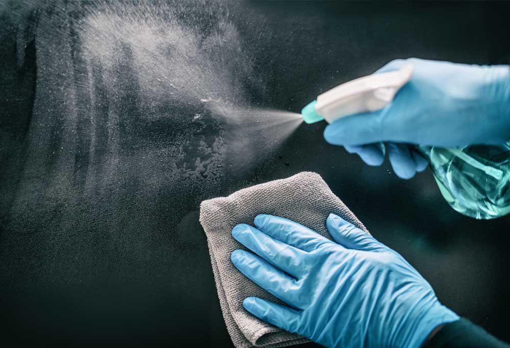 Set of hands with blue gloves spraying cleaner on dark glass and wiping it off with a grey cloth