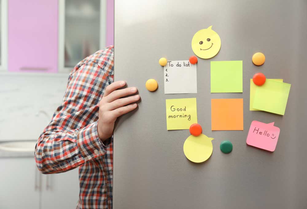 man in plaid shirt holding open fridge door with colorful notes and magnets on the door