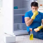 Man in blue shirt sitting on the floor in front of a fridge with cleaning spray, pinching his nose