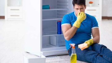 Man in blue shirt sitting on the floor in front of a fridge with cleaning spray, pinching his nose