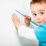 Toddler with wearing a teal shirt coloring on white wall with a teal colored pencil.