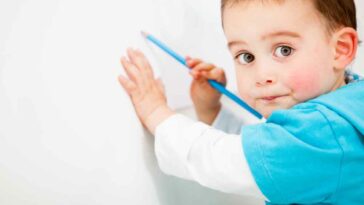 Toddler with wearing a teal shirt coloring on white wall with a teal colored pencil.