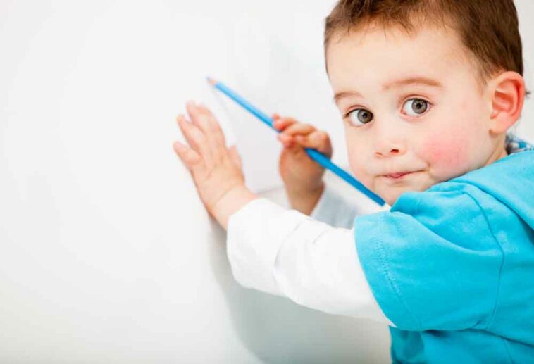 Toddler with wearing a teal shirt coloring on white wall with a teal colored pencil.