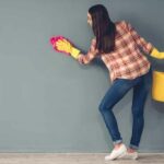 Woman in a plaid shirt and blue jeans scrubbing a blue/green wall with a sponge.
