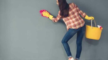 Woman in a plaid shirt and blue jeans scrubbing a blue/green wall with a sponge.