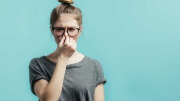 Woman with hair in a bun, wearing glasses, and holding her nose in front of a teal colored background