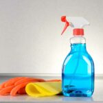 Bottle of glass cleaner next to a pair of gloves and a yellow rag on a grey surface.