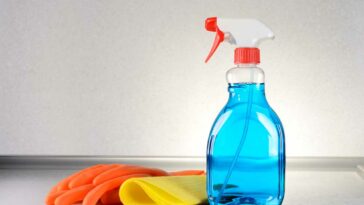 Bottle of glass cleaner next to a pair of gloves and a yellow rag on a grey surface.