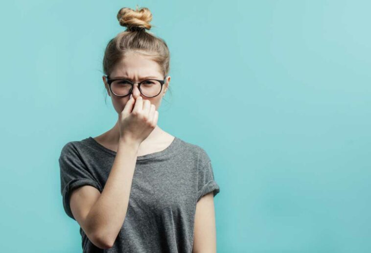 Woman with hair in a bun, wearing glasses, and holding her nose in front of a teal colored background