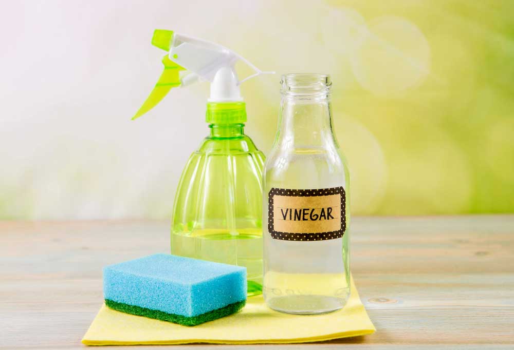 Glass bottle with a label that reads "Vinegar" , a spray bottle, and a blue sponge on a yellow cloth
