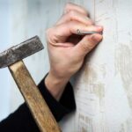 Hand holding a nail to a rustic wall with one hand while holding a hammer in the other.
