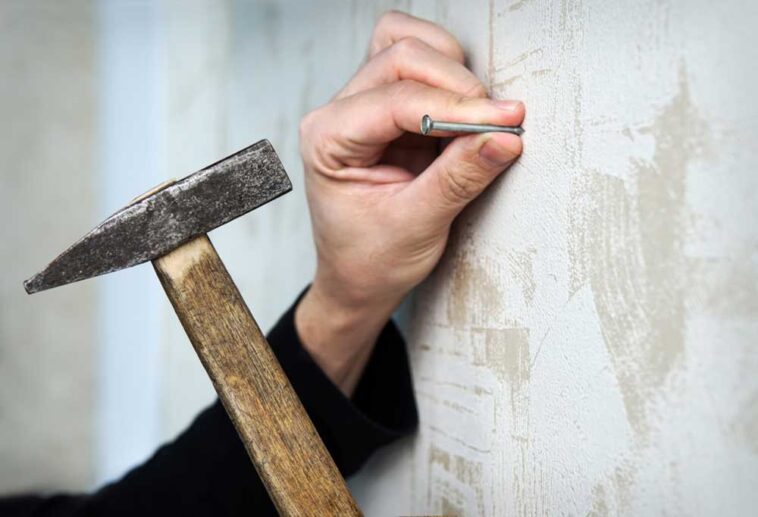 Hand holding a nail to a rustic wall with one hand while holding a hammer in the other.