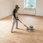 Person in overalls with an electric floor sander, sanding hard wood floors.