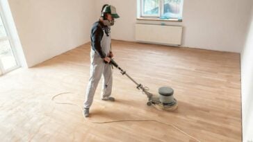 Person in overalls with an electric floor sander, sanding hard wood floors.