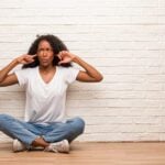 Woman sitting crossed legged on hardwood floors leaning against a white brick wall with her fingers in her ears.