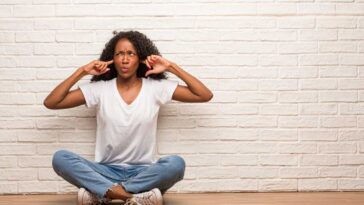 Woman sitting crossed legged on hardwood floors leaning against a white brick wall with her fingers in her ears.