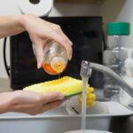Dish soap being poured on a sponge over a sink with water running and a microwave in the background.