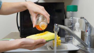 Dish soap being poured on a sponge over a sink with water running and a microwave in the background.
