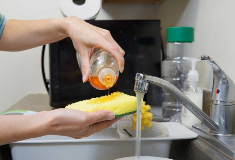 Dish soap being poured on a sponge over a sink with water running and a microwave in the background.