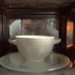 White bowl on a white plate, inside a microwave with a person looking in through the glass door.