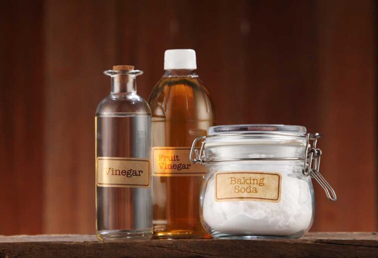 Bottles of vinegar and a glass jar of baking soda on a table with a dark wood background