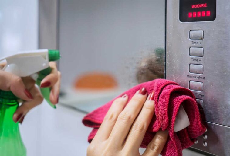 Hand holding rag and spray bottle, cleaning the front of a microwave