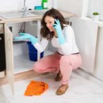 Woman kneeling in front of open under the sink cabinet with water on the floor, on the phone while cleaning up a leak