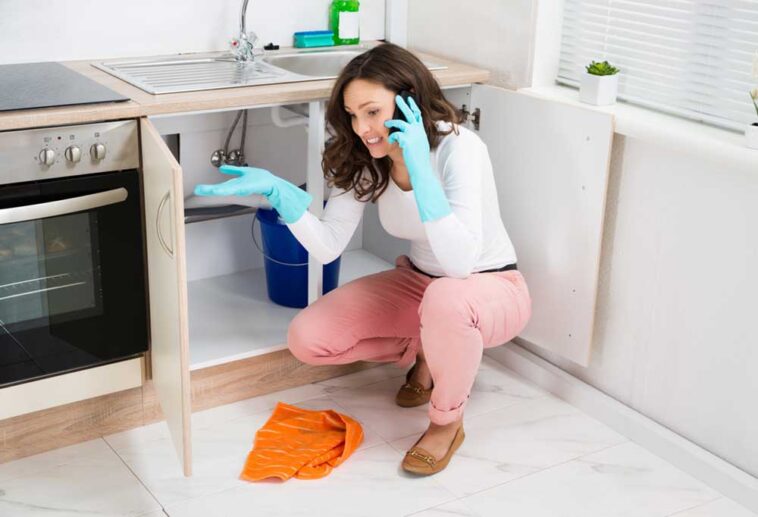 Woman kneeling in front of open under the sink cabinet with water on the floor, on the phone while cleaning up a leak