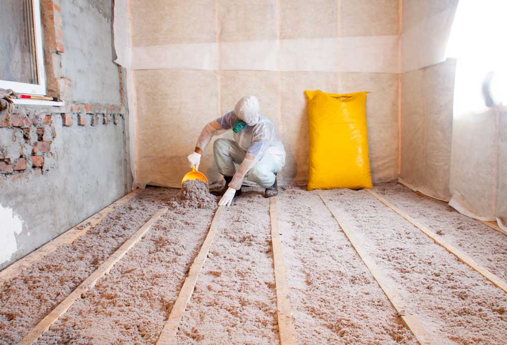 person wearing protective jump suit pouring insulation in between exposed floor joists.