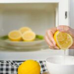 Hand squeezing a cut lemon into a bowl with other lemon halves in an open microwave in the background