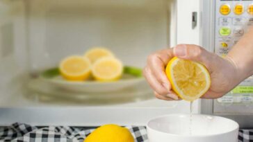 Hand squeezing a cut lemon into a bowl with other lemon halves in an open microwave in the background