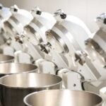 7 white KitchenAid mixers with heads tilted back lined up in a row