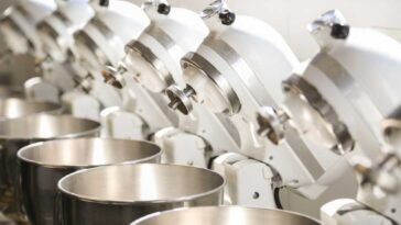 7 white KitchenAid mixers with heads tilted back lined up in a row