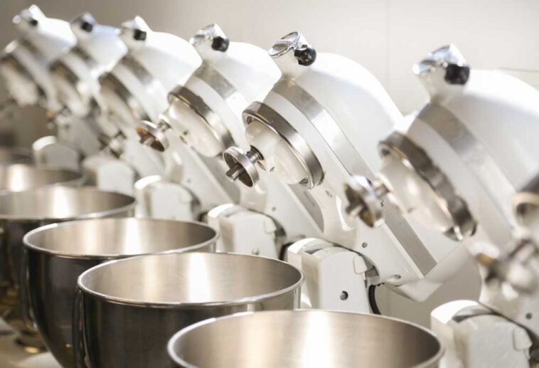 7 white KitchenAid mixers with heads tilted back lined up in a row