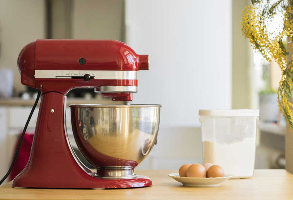 Red KitchenAid stand mixer on a kitchen counter with a bowl of eggs and a container of flower sitting next to it